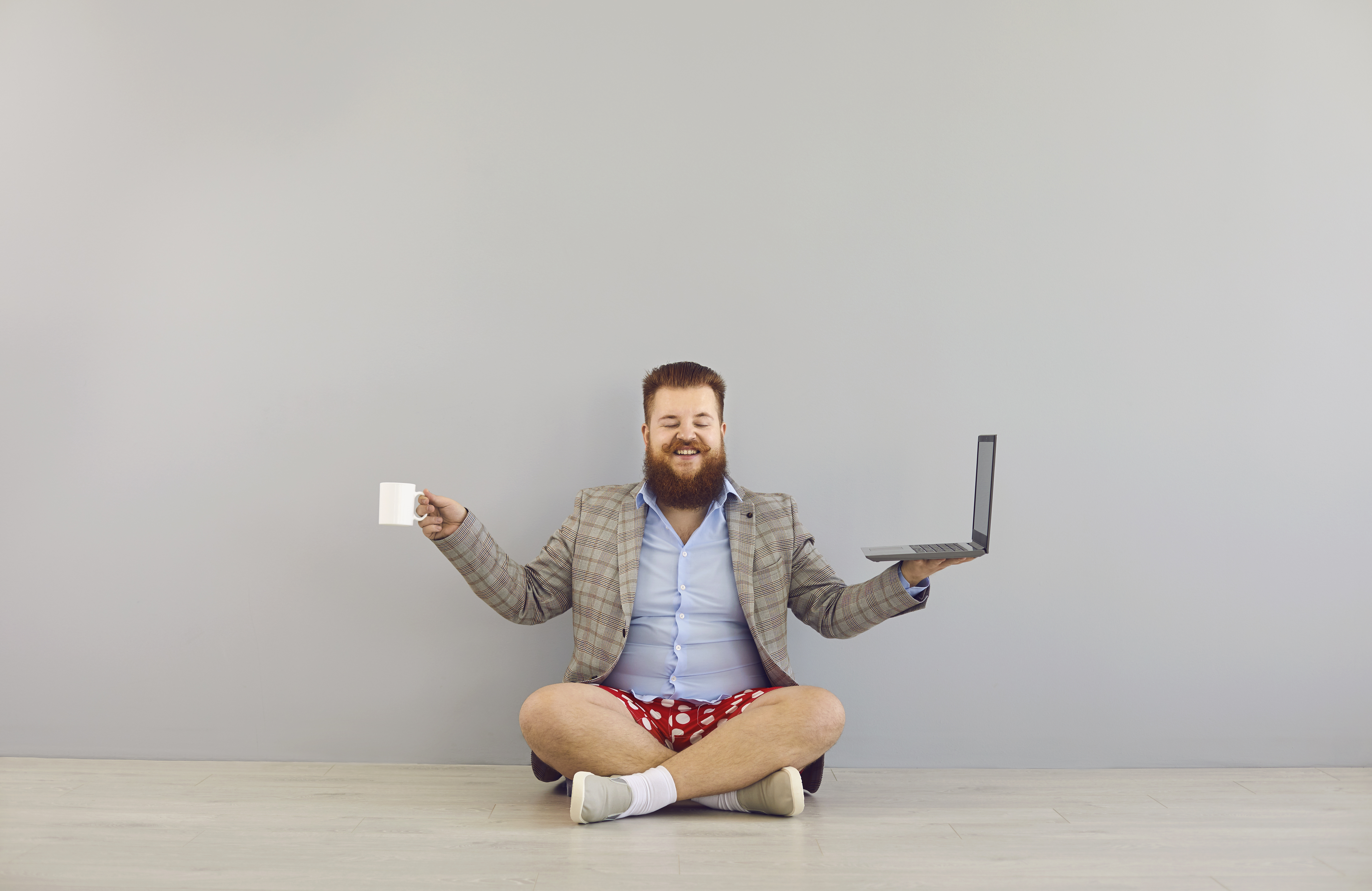Funny fat bearded man in a jacket and red shorts with a laptop on a gray background.Freelancer online distance work concept.